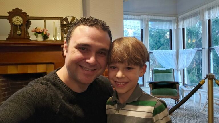 A man and a young boy smile for a selfie in a warmly lit room. Behind them is a wooden fireplace mantel with a clock, flowers, and a mirror. Large windows with lace curtains let in daylight, and a green chair sits nearby. A rope barrier is visible in the background.