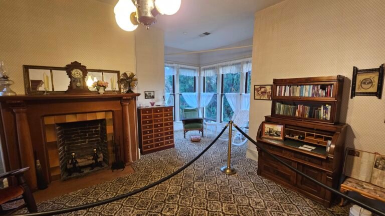Victorian-style room with patterned carpet, wooden fireplace mantel, vintage clock, and chest of drawers. Bookshelves and a writing desk are on the right, a roped barrier in front. Sunroom with lace curtains and rocking chair visible through large windows.