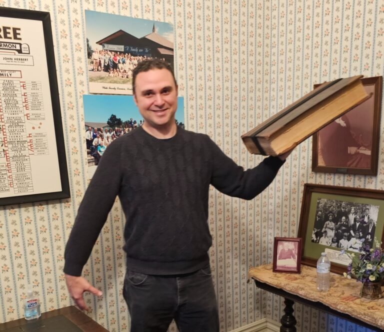 A smiling man in a dark sweater stands indoors holding a large, thick book with yellowed pages. Behind him are framed photos, a water bottle on a table, and wallpaper with a small floral pattern. Family memorabilia decorates the room.