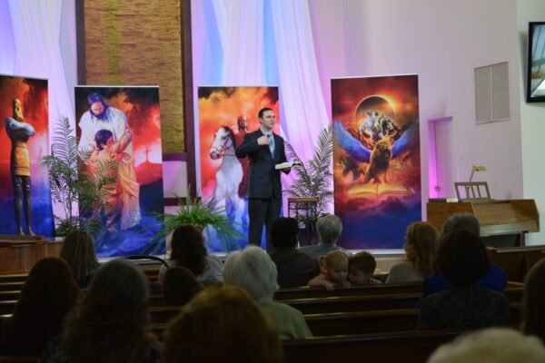 A man in a suit speaks, holding "The Least of the Least: From Crime to Christ" by Wyatt Allen, at the front of a church with seated attendees. Colorful biblical banners and potted ferns decorate the stage lit by streaming light.