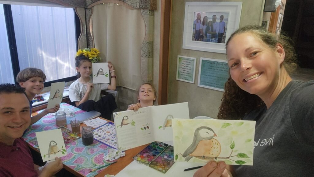 A smiling woman takes a selfie with three children and a man at a table. They all hold up watercolor bird paintings. Art supplies and a book are on the table. Behind them are framed photos, a vase of yellow flowers, and colorful posters on the wall.