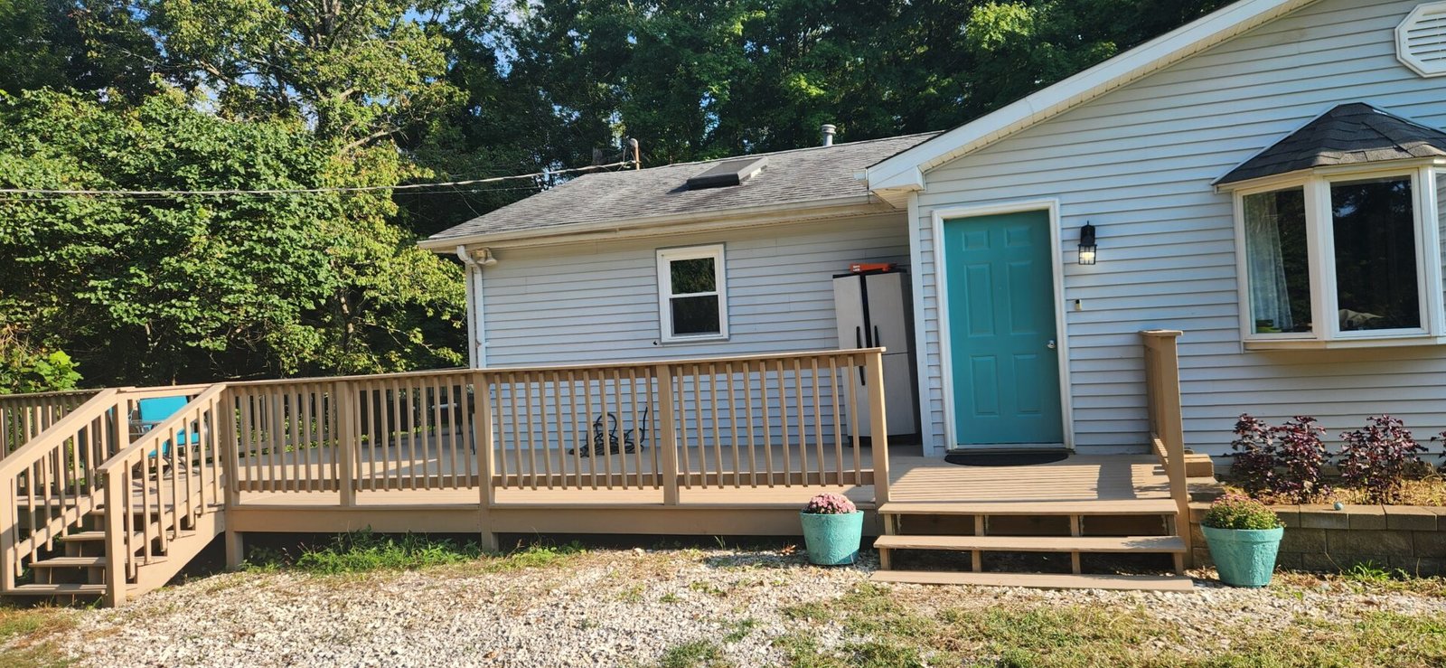 A light gray house with a turquoise front door and bay window sits next to a wooden ramp and stairs with tan railings. Two blue planters with flowers are near the steps. Trees fill the background, and gravel covers the ground in front.