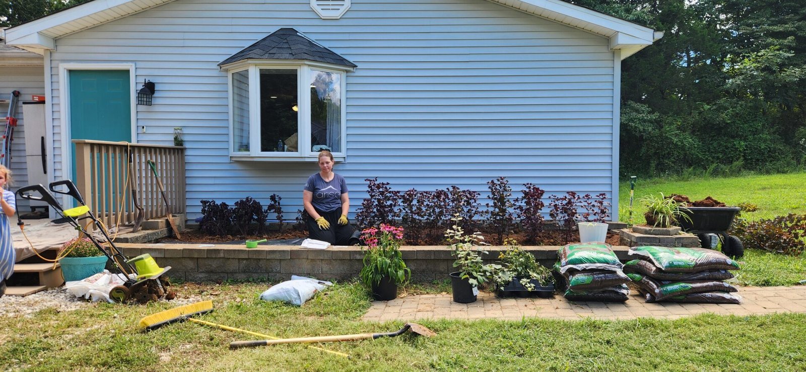 A person kneels and smiles in a flower bed in front of a light blue house. Gardening tools, soil bags, mulch, and potted plants are scattered around, suggesting ongoing landscaping work. Bushes are planted along the house, and a lawn surrounds the scene.