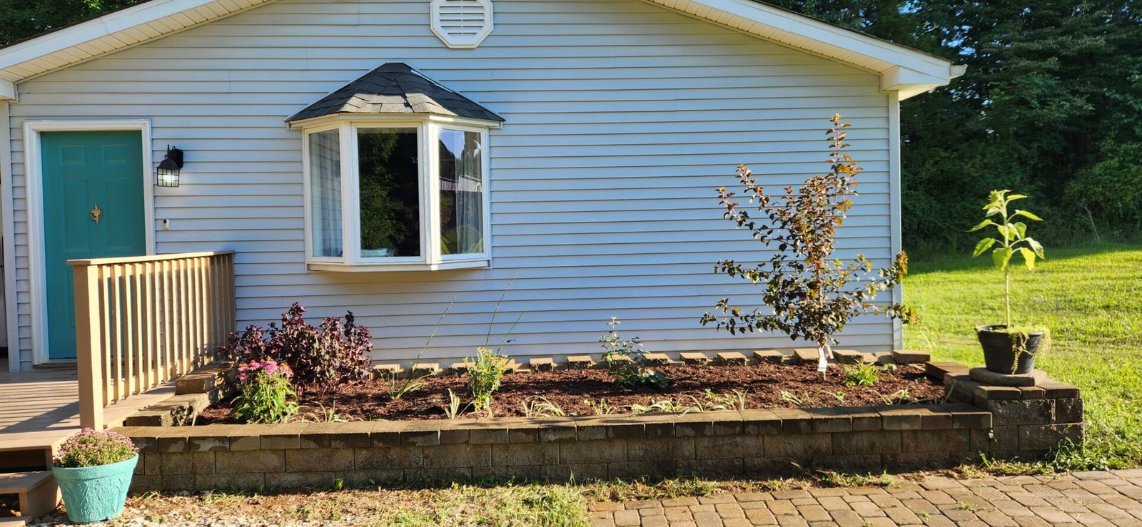 A small raised garden bed with flowers and small shrubs sits in front of a light blue house with white trim. A teal door and a bay window are visible. Two large potted plants are placed at each end of the garden, and a brick pathway runs in front.