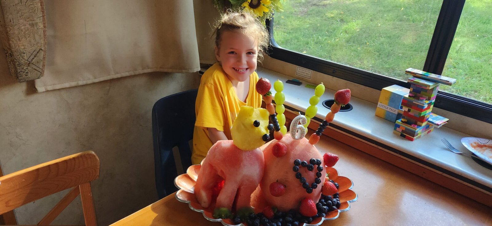 A smiling girl in a yellow shirt sits indoors beside a camel-shaped fruit sculpture made of watermelon, cantaloupe, strawberries, blueberries, and skewered fruit. The sculpture sits on a platter; sunlight streams through a window behind her. A Jenga game is nearby.