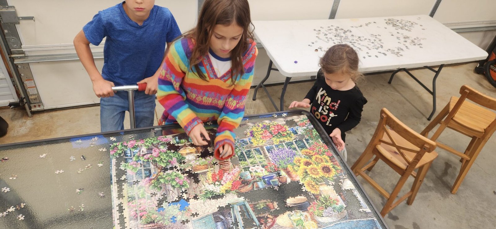Three children work together on a large floral jigsaw puzzle on a glass table in a garage. The oldest girl wears a colorful striped sweater, a boy in blue stands beside her, and a younger girl in black stands nearby. Another table with loose pieces is in the background.