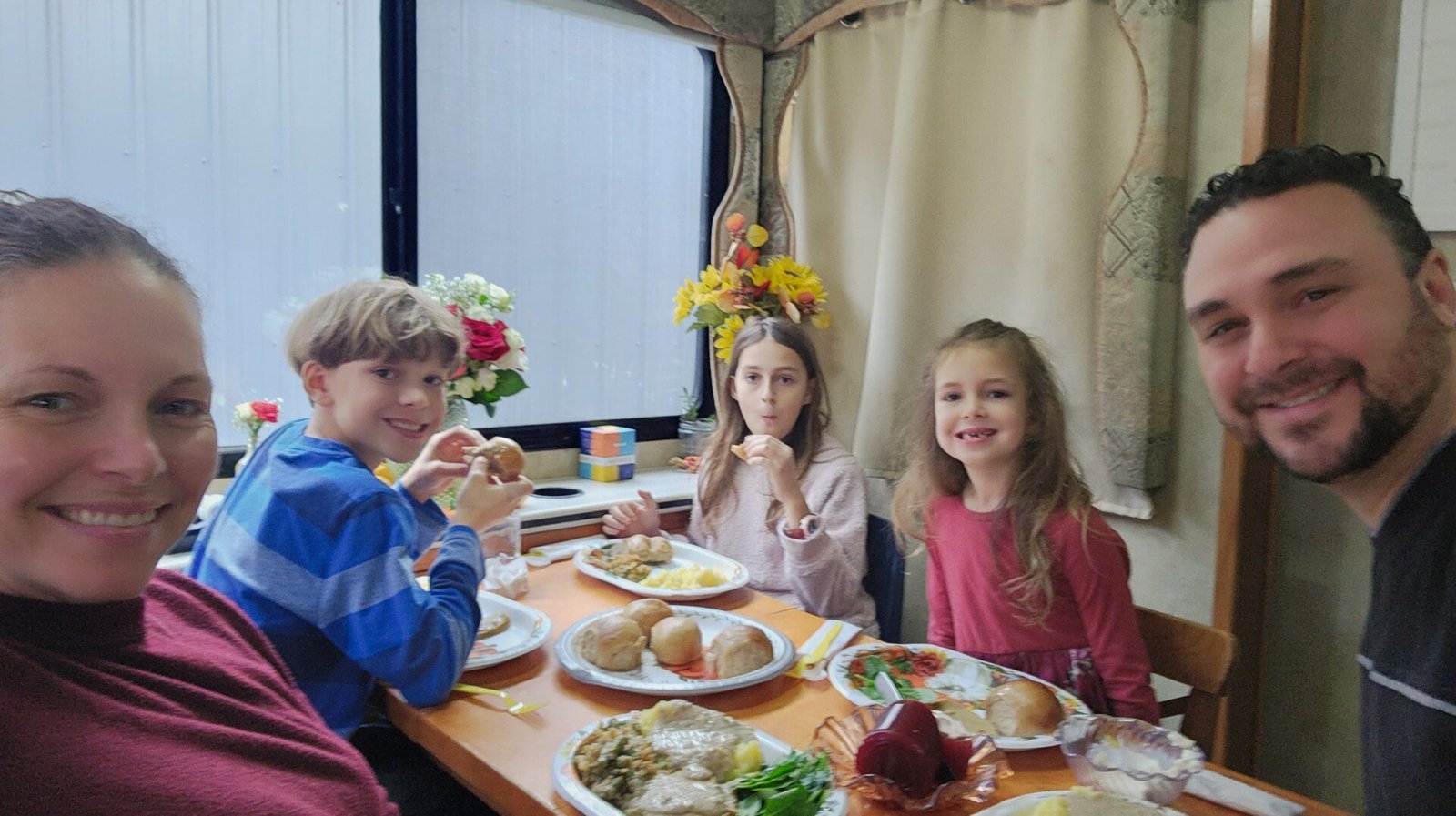 A smiling family of five sits around a table by a window, sharing a meal. The table is set with plates of food, including rolls, potatoes, and vegetables. The background features flowers, curtains, and a cozy indoor setting.