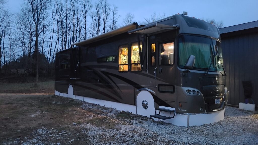 A large, dark gray RV with slide-outs is parked on gravel near a gray building at dusk. The RVs interior is warmly lit and its windows glow. Leafless trees and a cloudy sky are in the background, and white skirting surrounds the RV&rsquo;s base.