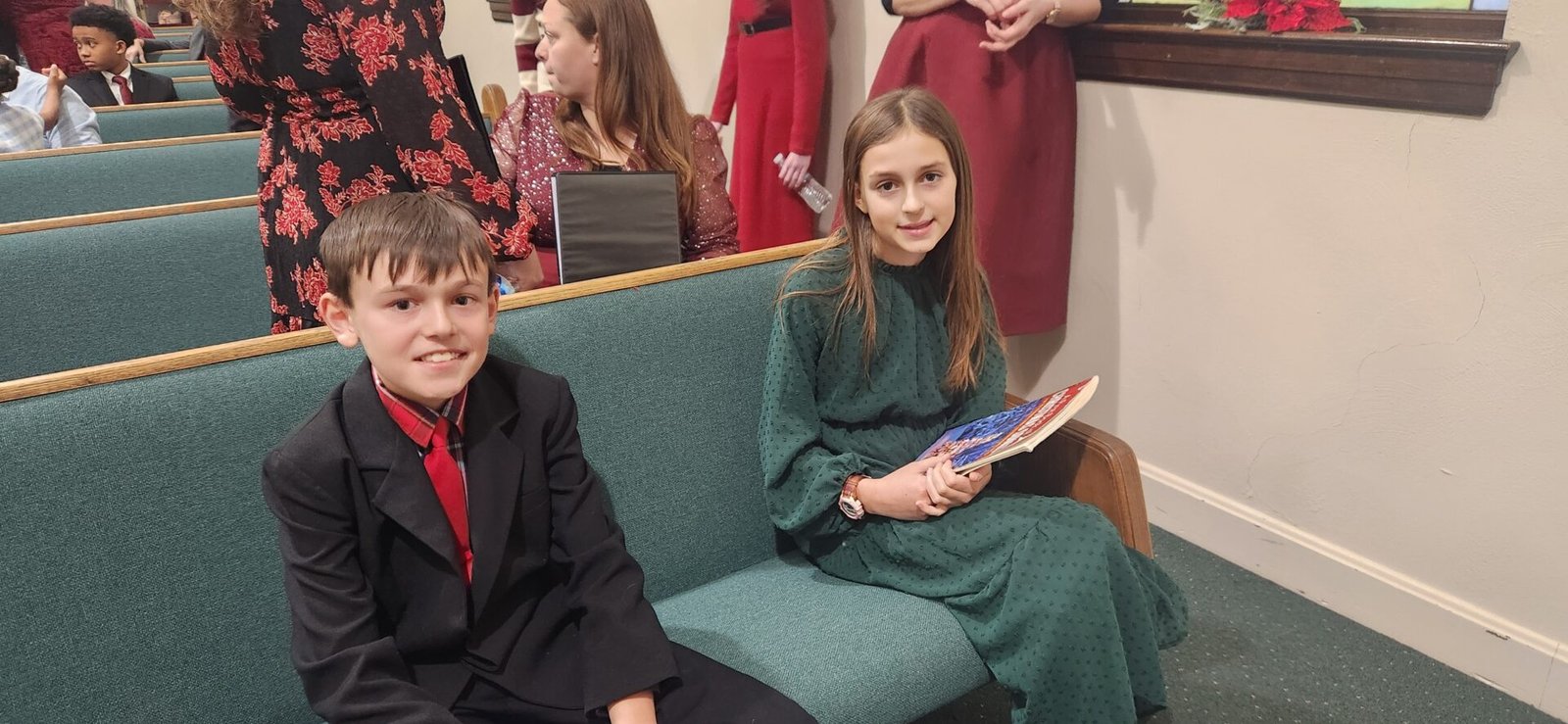 A boy in a black suit with a red tie and a girl in a green dress holding a book sit on a church pew. Other children in formal clothes sit and stand nearby, with holiday decorations visible in the background.